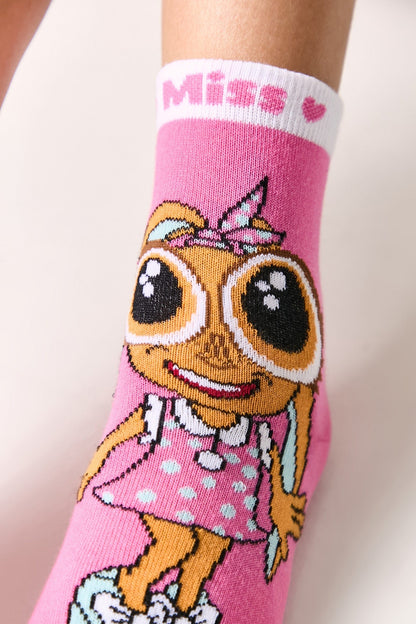 A pink ankle sock features a cartoon girl with large eyes and a polka-dot dress, with the word Miss and a small heart printed on the white ribbed cuff.