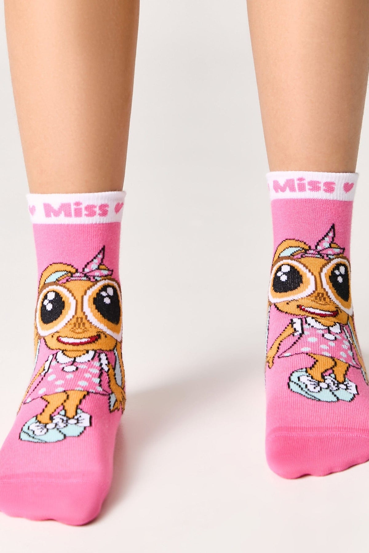 A pair of pink crew socks worn on a person's feet, featuring a cartoon character with large eyes, a polka-dot dress, and the word "Miss" on the white ribbed cuffs.