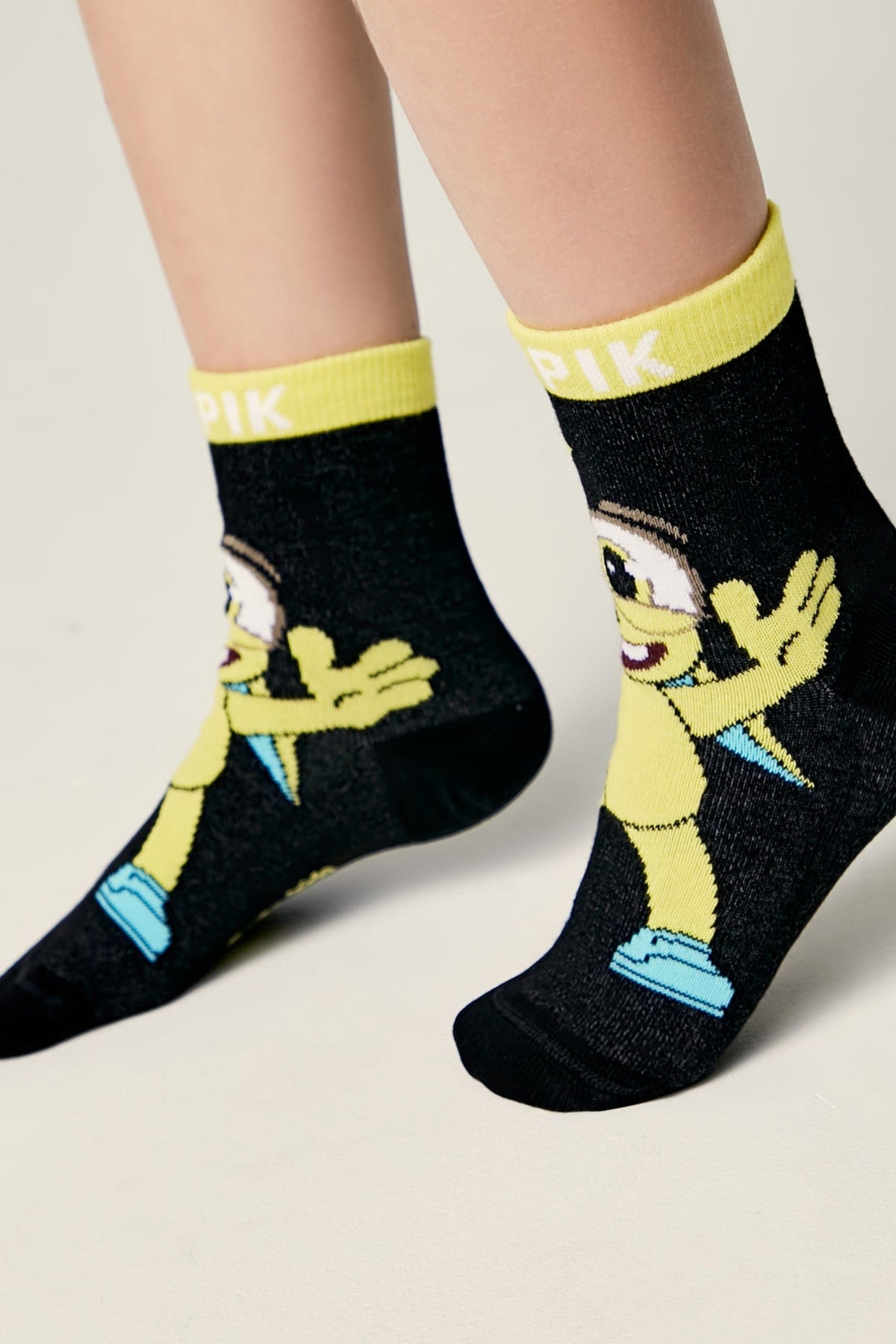A pair of black ankle socks featuring a cartoon character with a single eye, yellow skin, and blue shoes, finished with a bright yellow ribbed cuff.