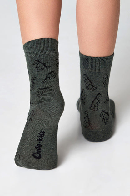 A pair of dark green crew socks on a child's feet, featuring a repeating pattern of black dinosaur outlines and the text Cute kids printed on the soles.
