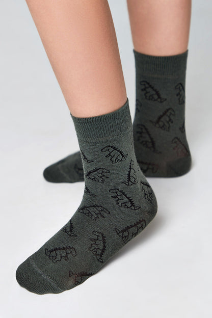 Olive green crew-length socks featuring a repeating dark brown dinosaur pattern worn on a person's lower legs against a plain background.