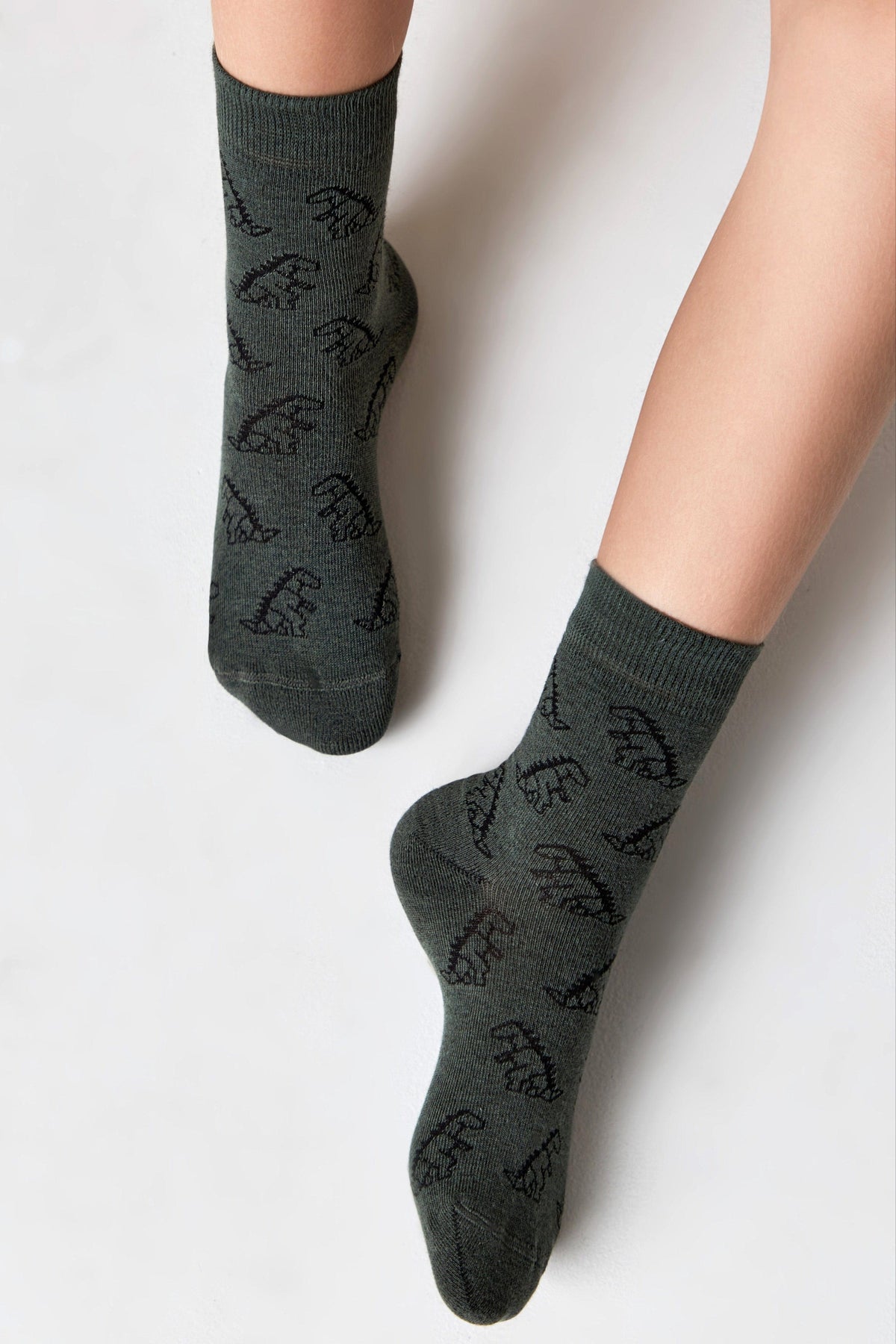 Dark green crew socks featuring a repeating pattern of black dinosaur outlines, worn on a person's legs against a white background.