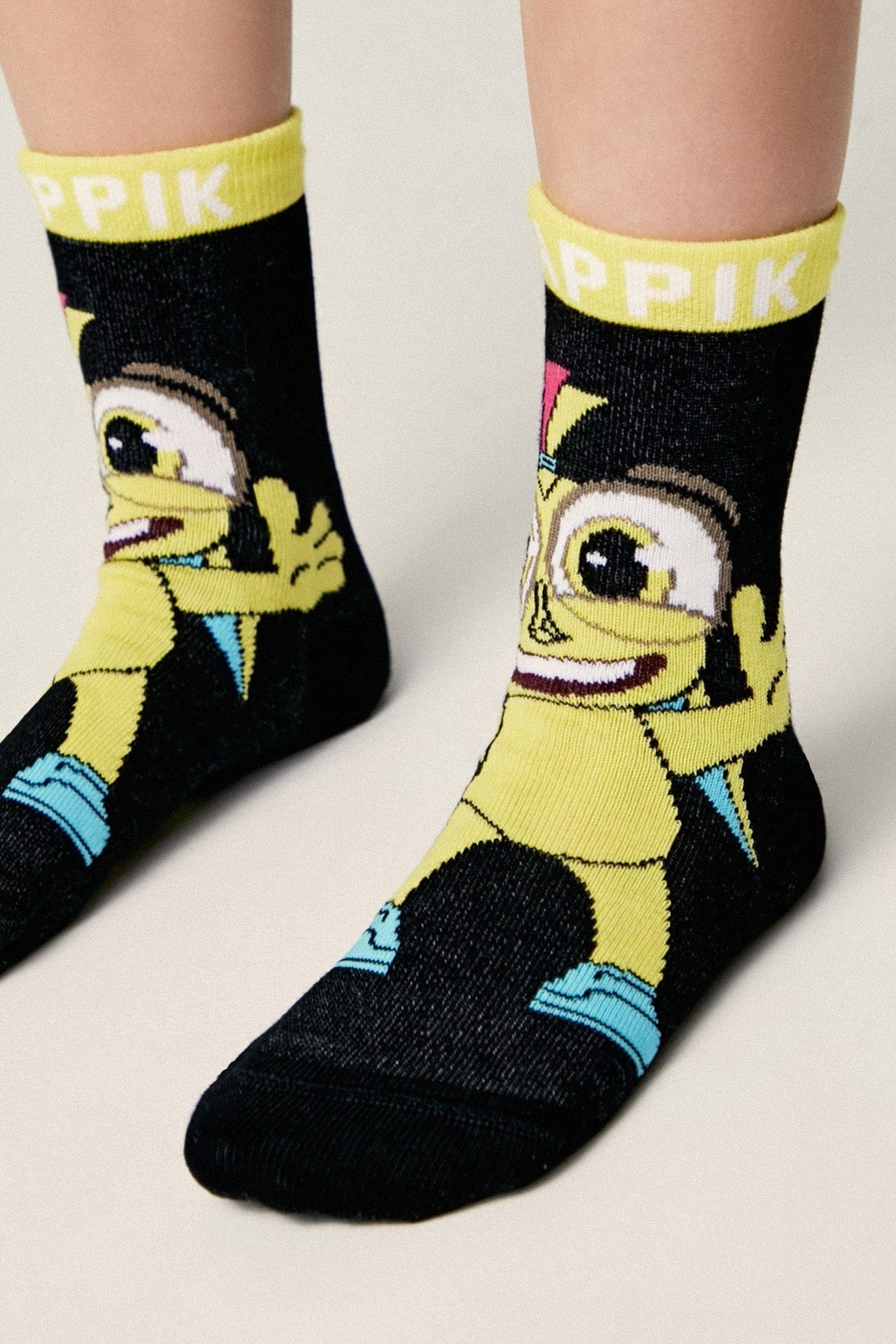 A pair of black ankle socks featuring a cartoon character with yellow skin and blue shoes, with the word PIK printed in white on the yellow cuffs.