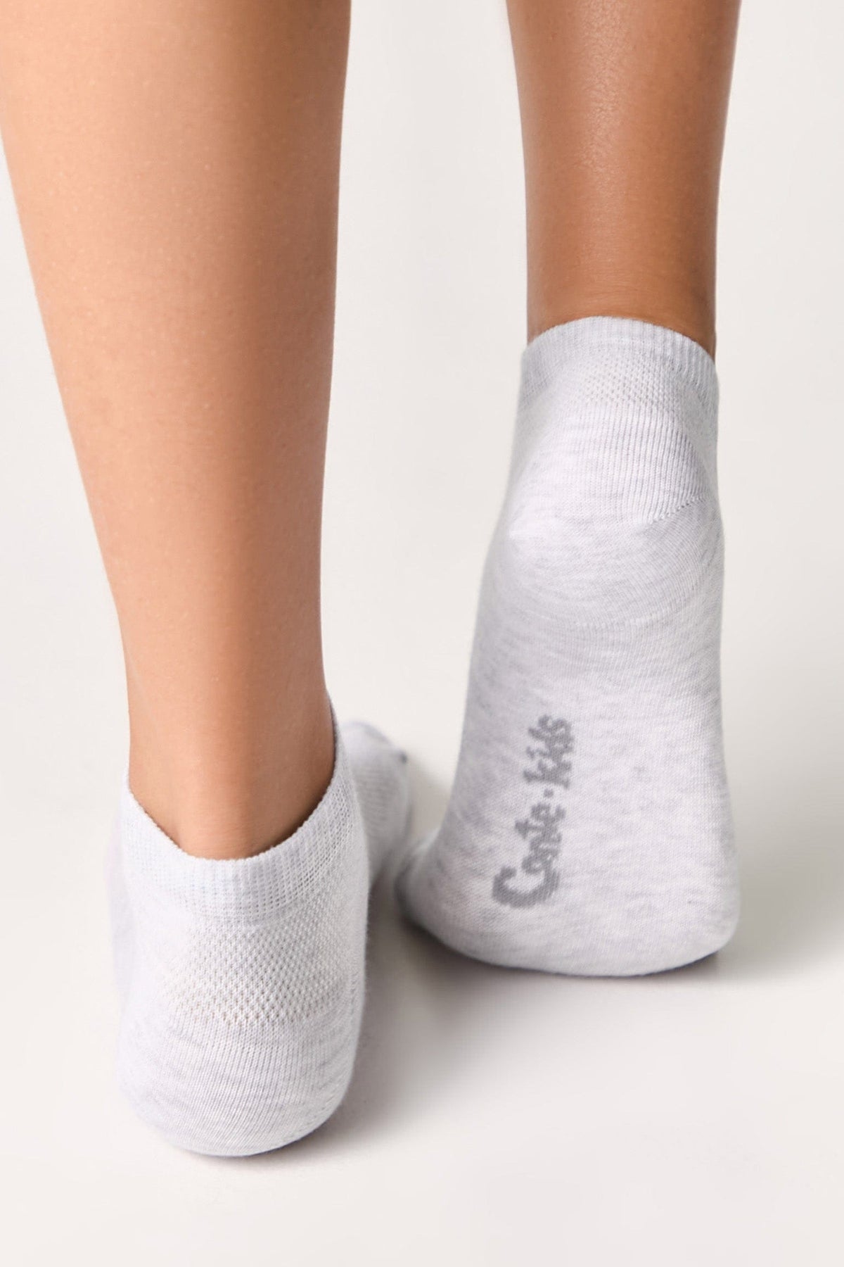 A pair of light grey, ankle-length socks worn on a person's feet, showing the back of the heels with a logo printed on the sole of the right sock.