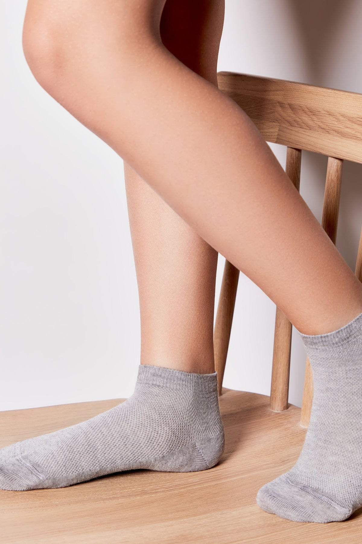 A pair of grey ankle socks on feet resting on a light wood chair.