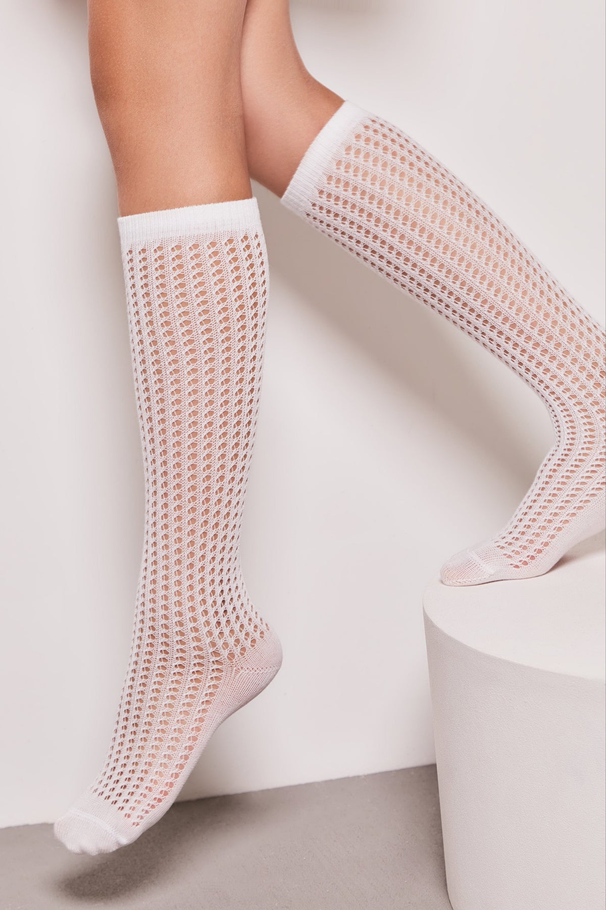 A pair of white, knee-high, open-knit patterned socks worn on a person's legs against a plain background with a white cylindrical stand.