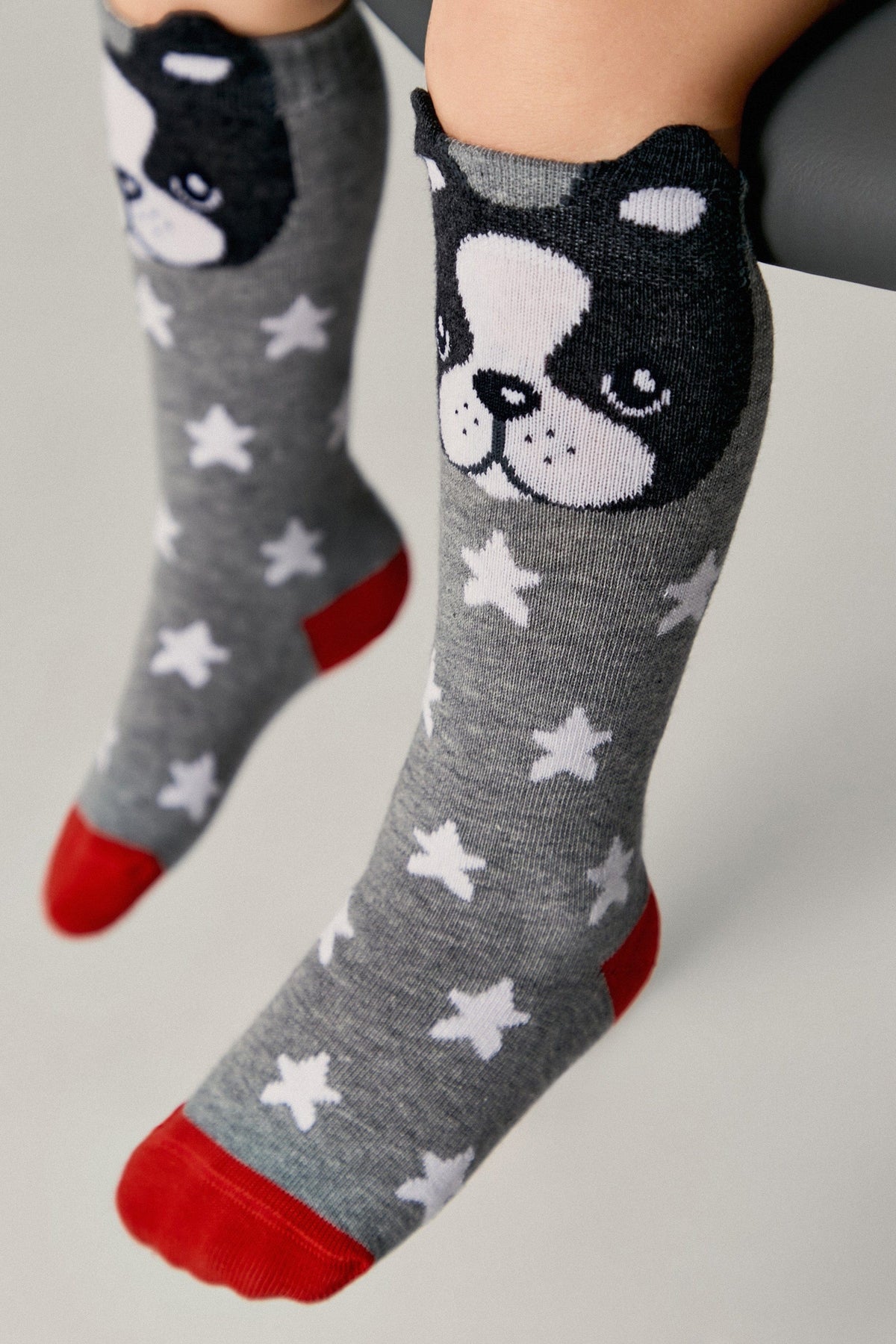 Gray knee-high socks featuring a white star pattern and a black and white puppy face at the top, accented with a bright red heel and toe.