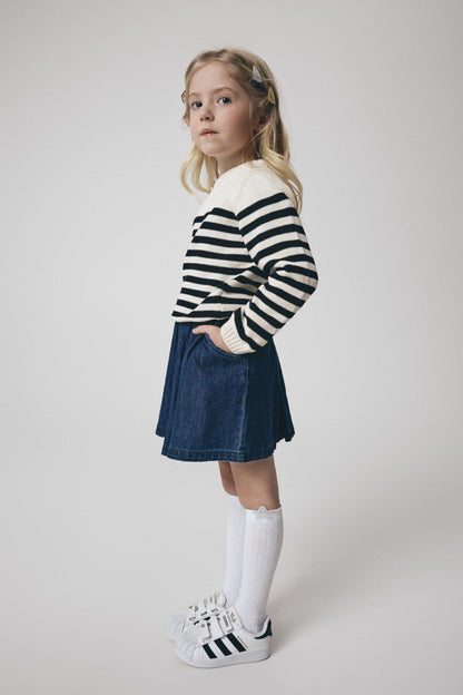 A young girl stands in profile wearing a cream-colored sweater with horizontal black stripes, a dark blue denim skirt, white socks, and white sneakers.