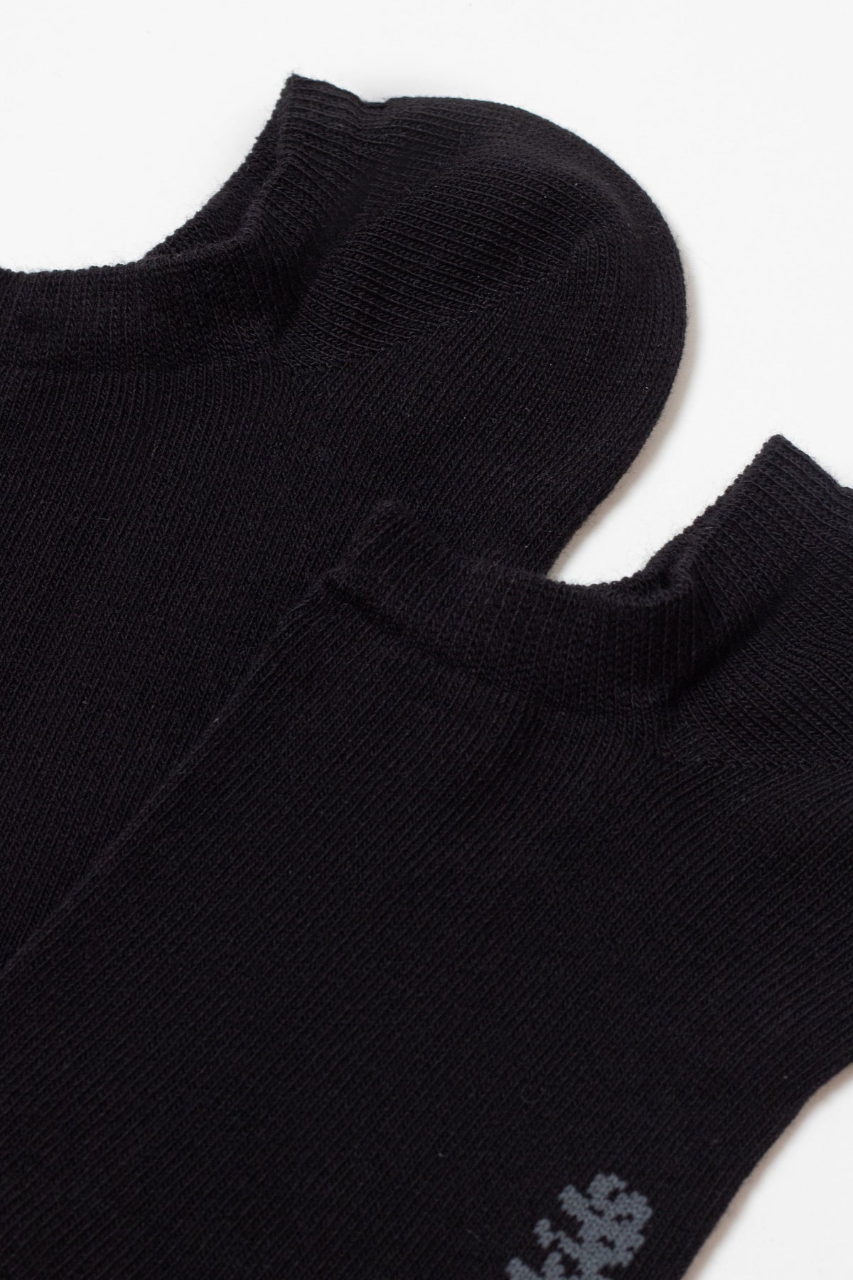 Two black low-cut ankle socks are folded and overlapping against a plain white background, showing their ribbed cuffs and a small gray detail near the edge of one sock.