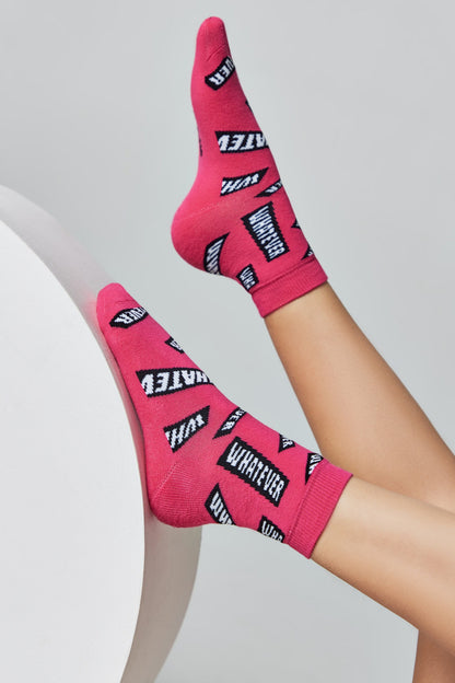 Bright pink ankle socks featuring a repeating black and white pattern of the word WHATEVER, worn on the feet of a person leaning against a white rounded surface.
