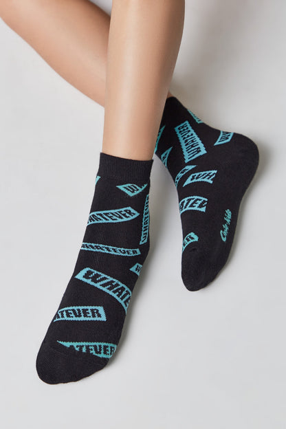 A pair of black ankle socks featuring a repeating cyan pattern of the word WHATEVER in a rectangular font style worn on a person's feet.