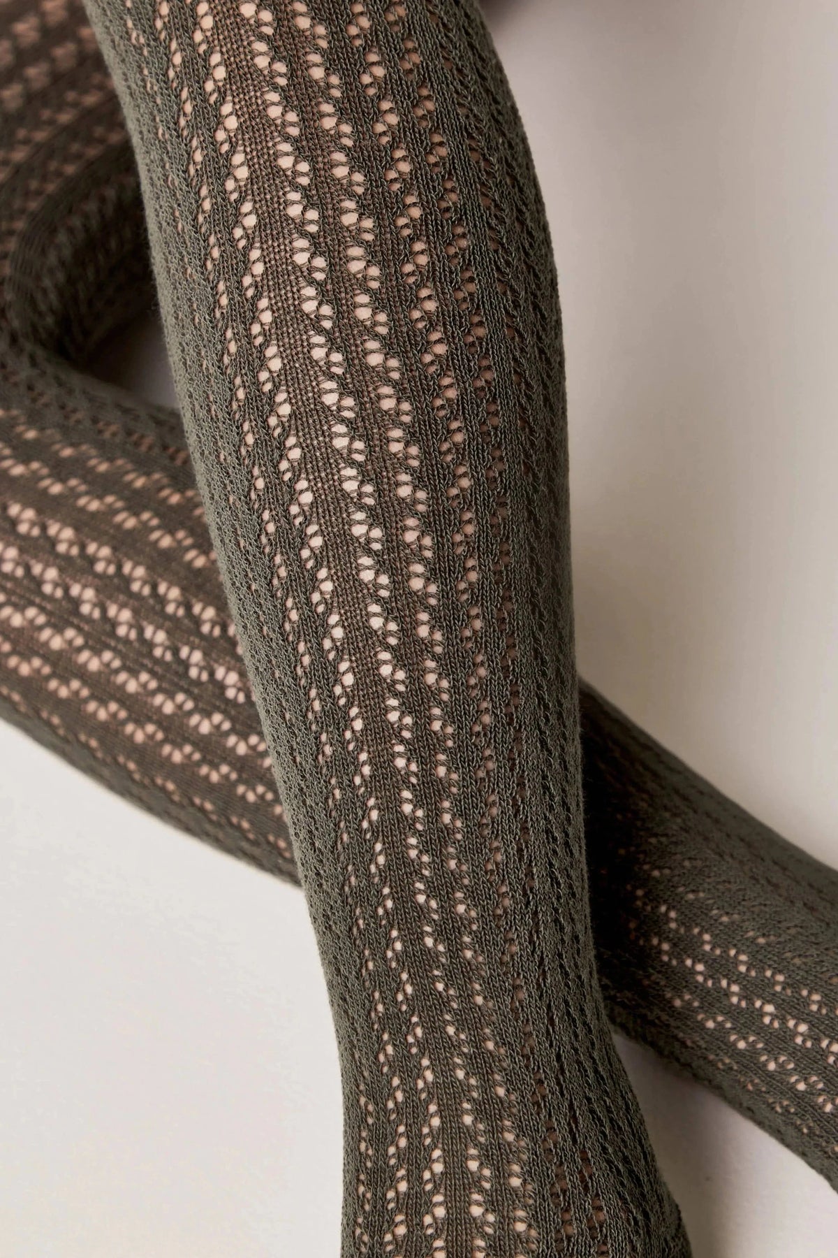 A close-up of olive green tights featuring an intricate vertical pointelle knit pattern with small, openwork geometric cutouts throughout.