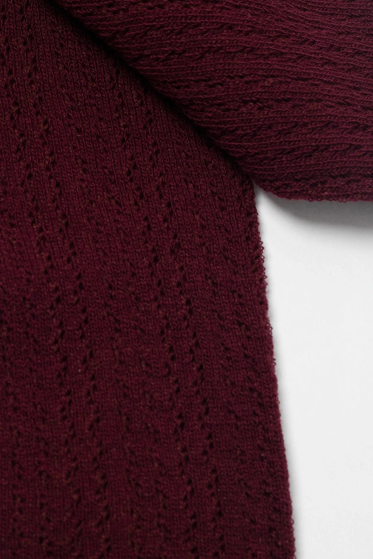 A close-up of a burgundy knit fabric featuring vertical, eyelet lace-style detailing against a white background.