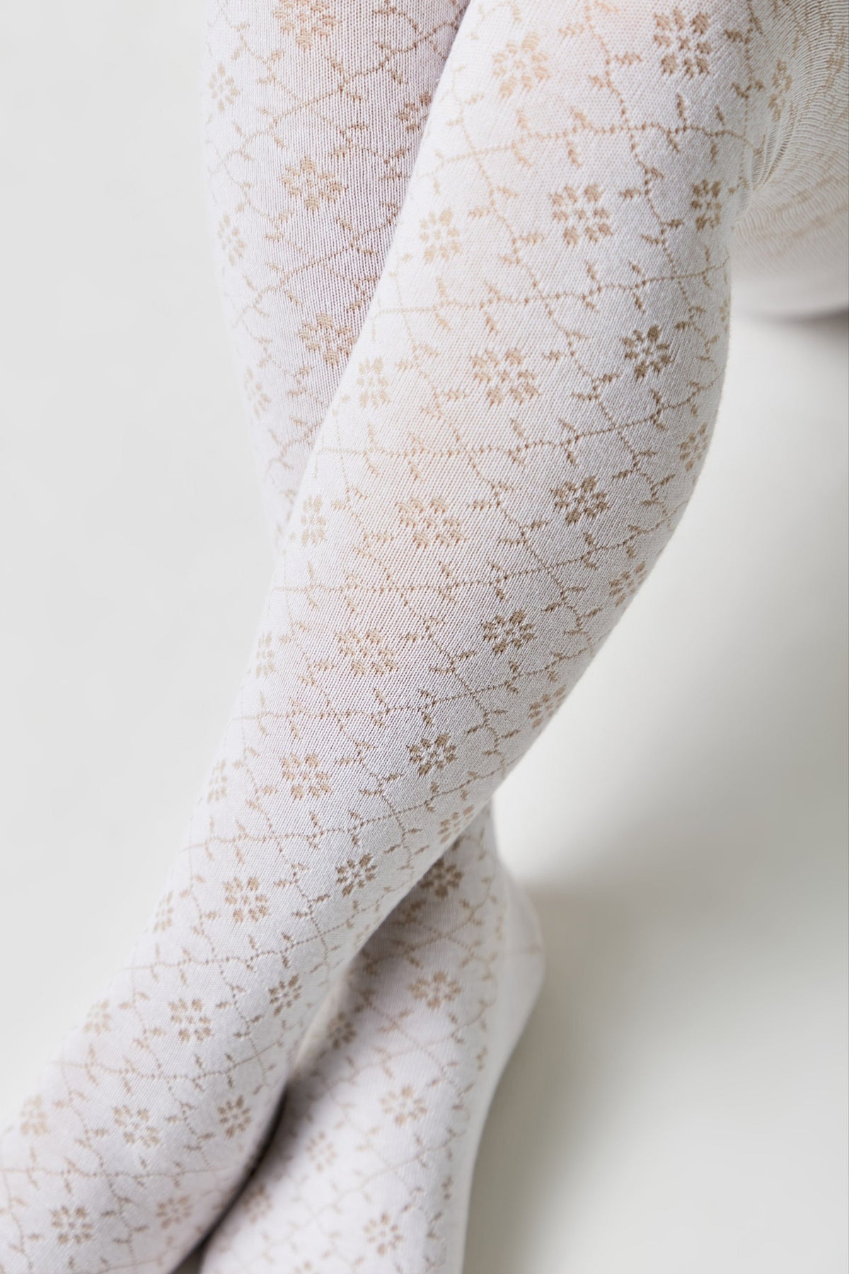 White tights featuring a repeating diamond grid pattern filled with small beige floral or snowflake motifs, worn on crossed legs against a plain background.