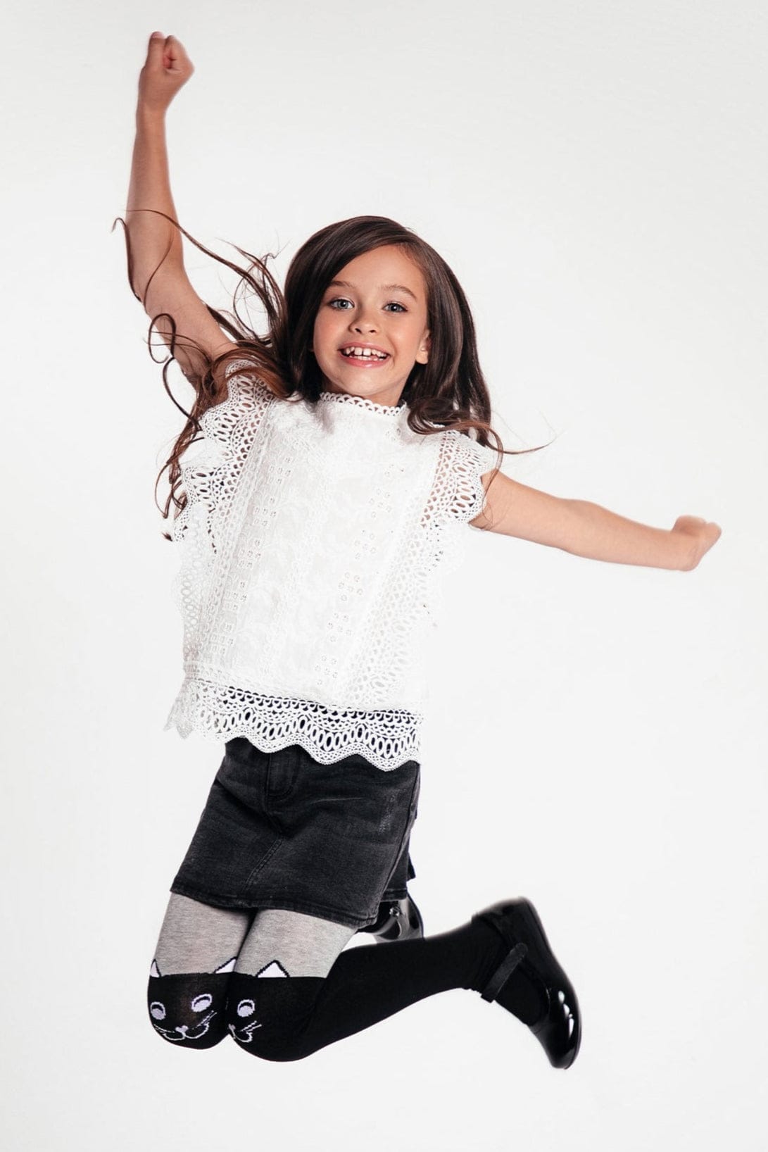 A young girl with long brown hair jumps in the air wearing a white sleeveless lace top, a black denim skirt, and grey tights with cat face designs on the knees.
