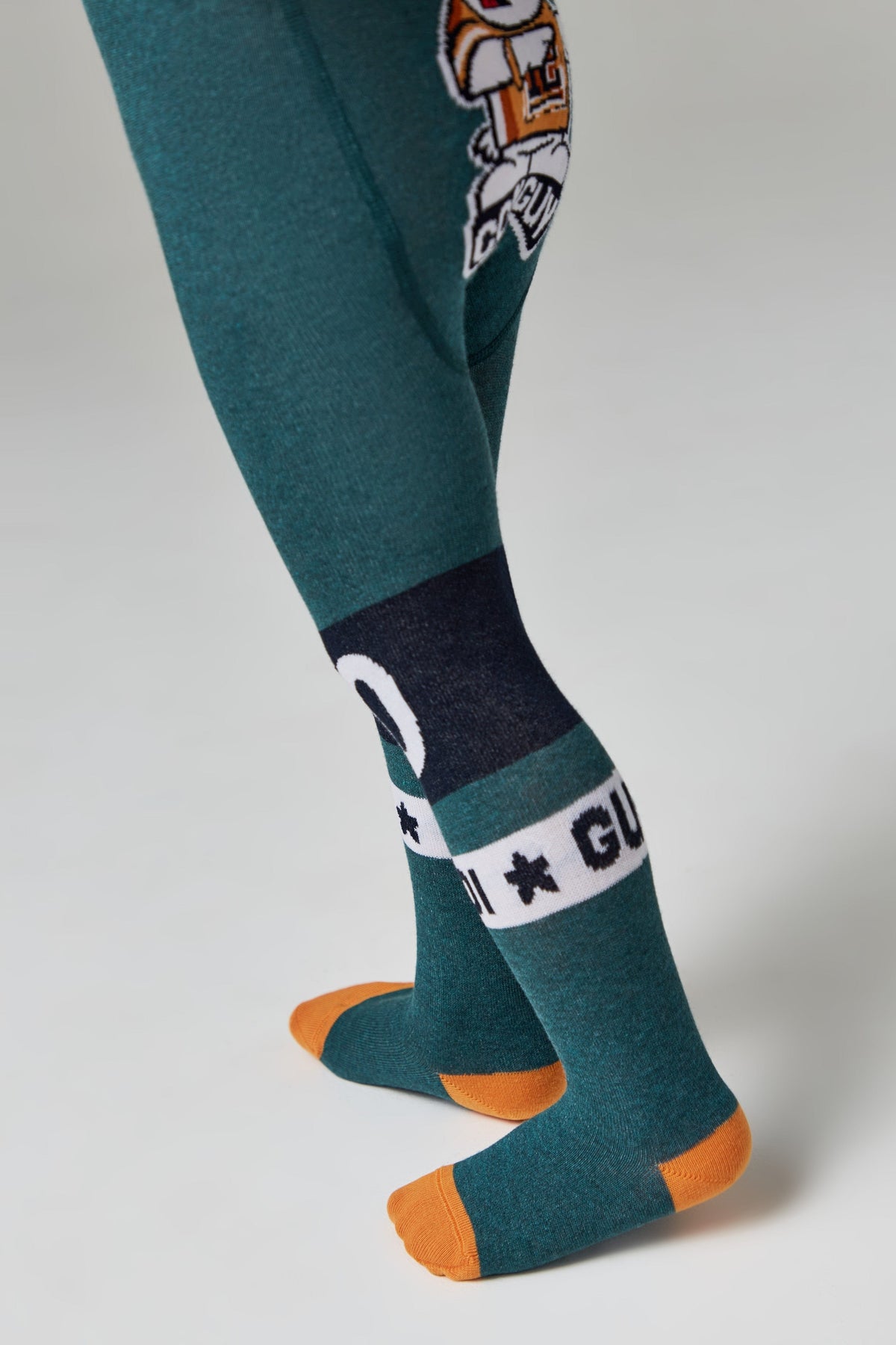 A child's legs are shown wearing teal tights that feature an orange heel and toe, with a horizontal dark blue band above the ankle containing white text and star patterns.