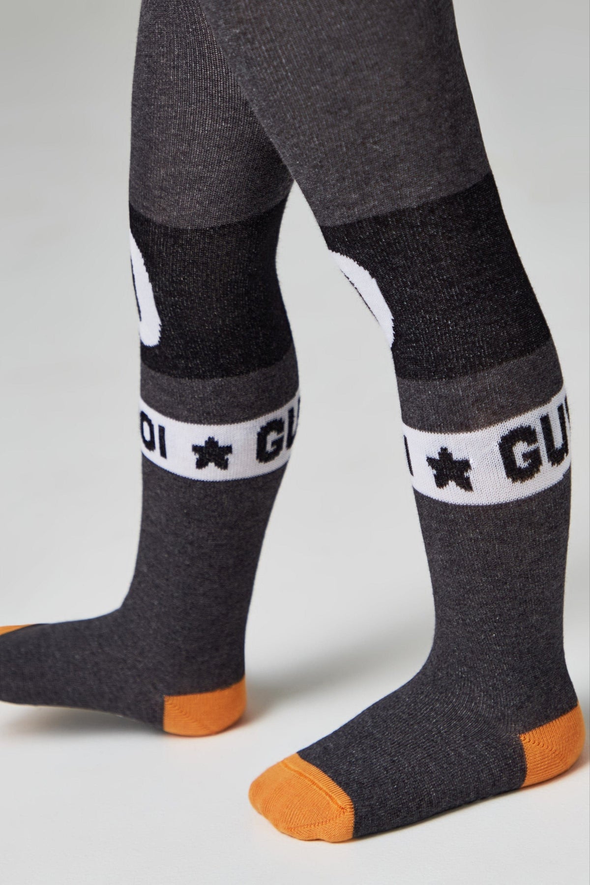 A pair of children's grey knit tights featuring black and white horizontal bands across the shins with white star and letter detailing, finished with contrasting orange toe sections.