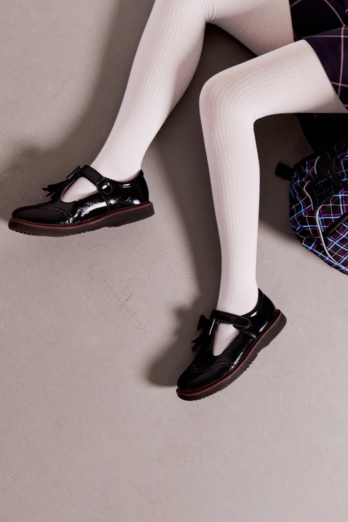 Black patent leather T-strap shoes with tassels and brogue detailing worn with opaque white tights on a grey floor.