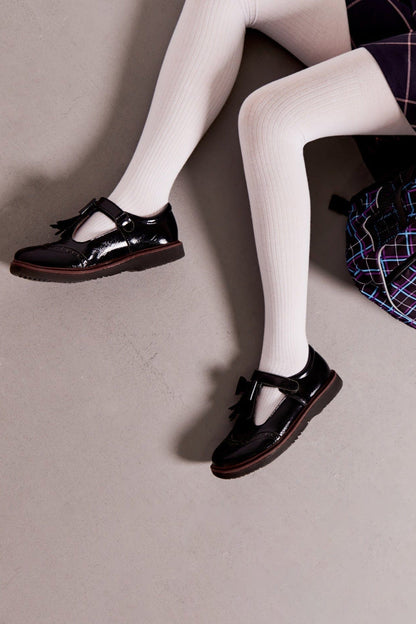 Black patent leather T-strap shoes with tassels and brogue detailing worn with opaque white tights on a grey floor.