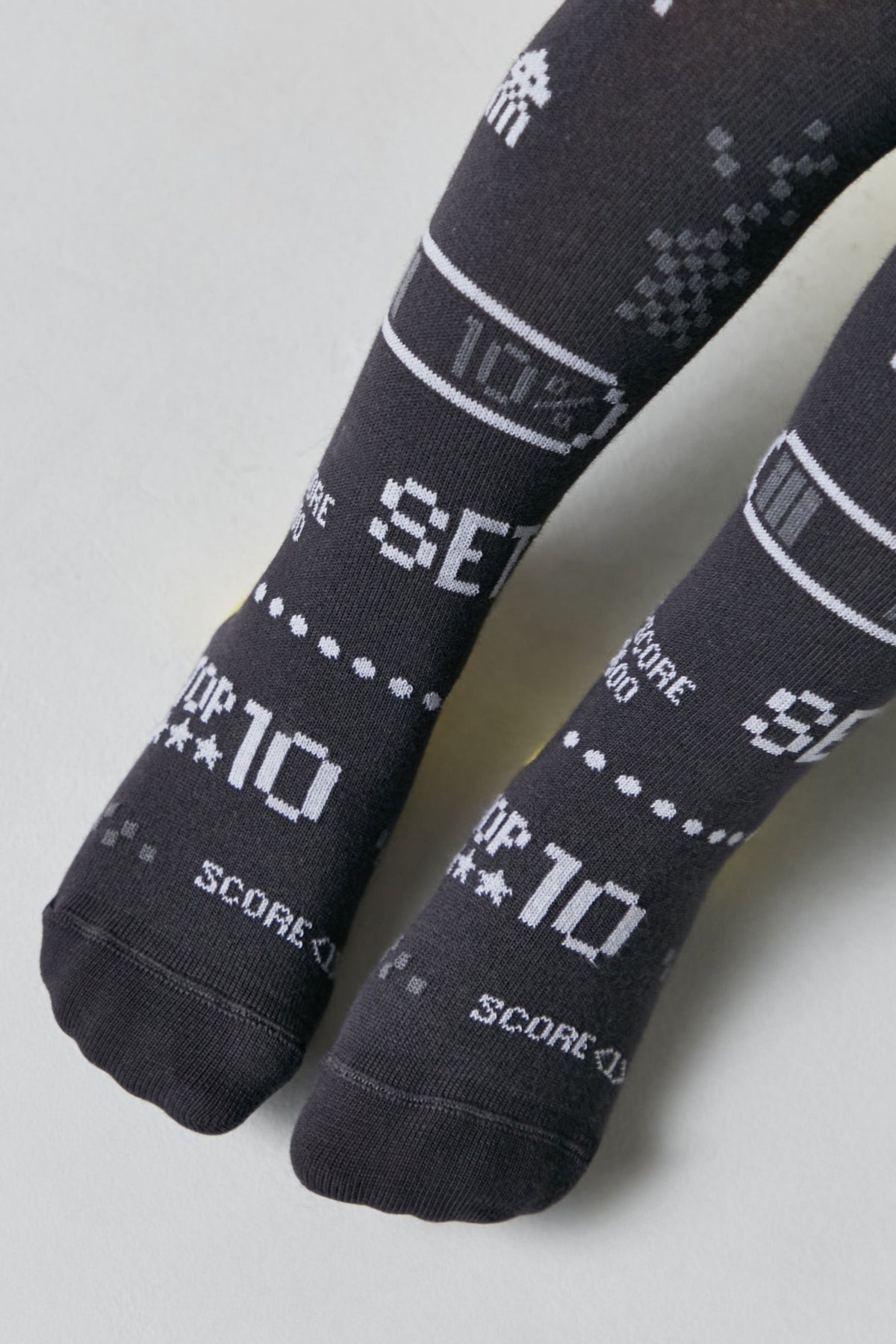 A pair of dark grey children's socks featuring a white pixelated video game-inspired design with graphics including space invaders, battery icons, and text that says top 10 and score.