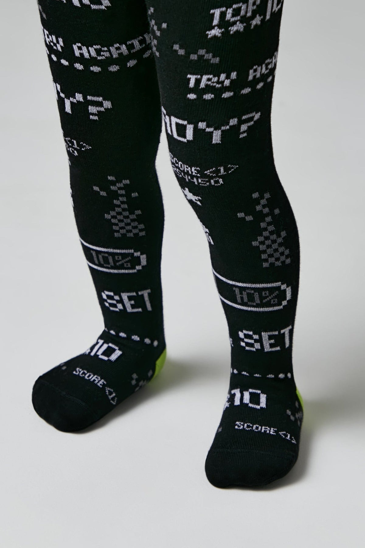 Black tights for children featuring a pixelated, retro video game-inspired pattern in white including text like TRY AGAIN, 10 PERCENT, and SCORE along with battery icons.