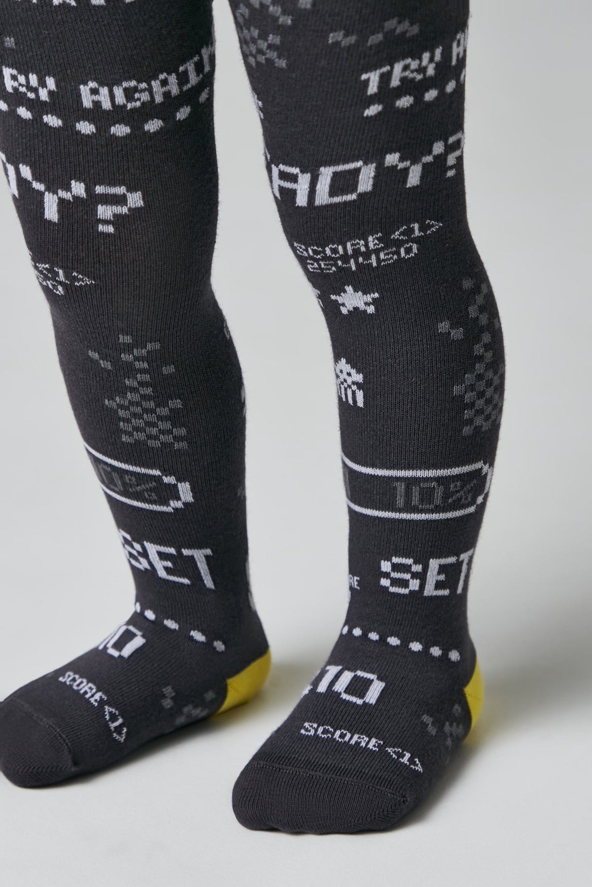 A pair of dark grey knitted tights for children featuring a white 8-bit retro video game pattern with text including "TRY AGAIN", "READY?", "SCORE <1>", and "SET" along with battery icons.