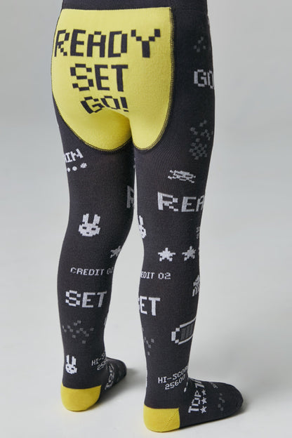 A pair of children's dark gray leggings featuring a yellow seat panel with the pixelated black text 'READY SET GO!' and various white retro arcade-style icons and text patterns printed throughout the legs.