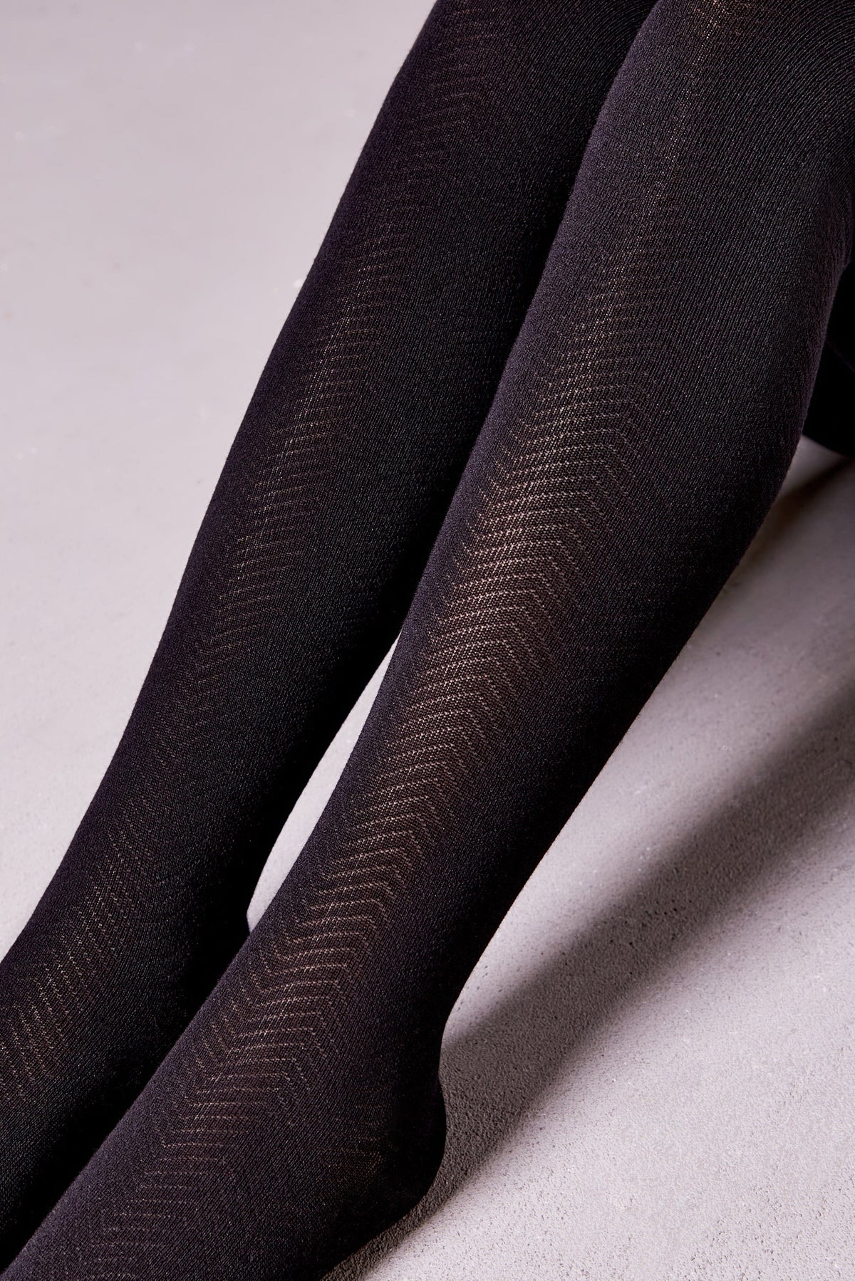 A close-up view of legs wearing black opaque tights featuring a subtle textured herringbone pattern.