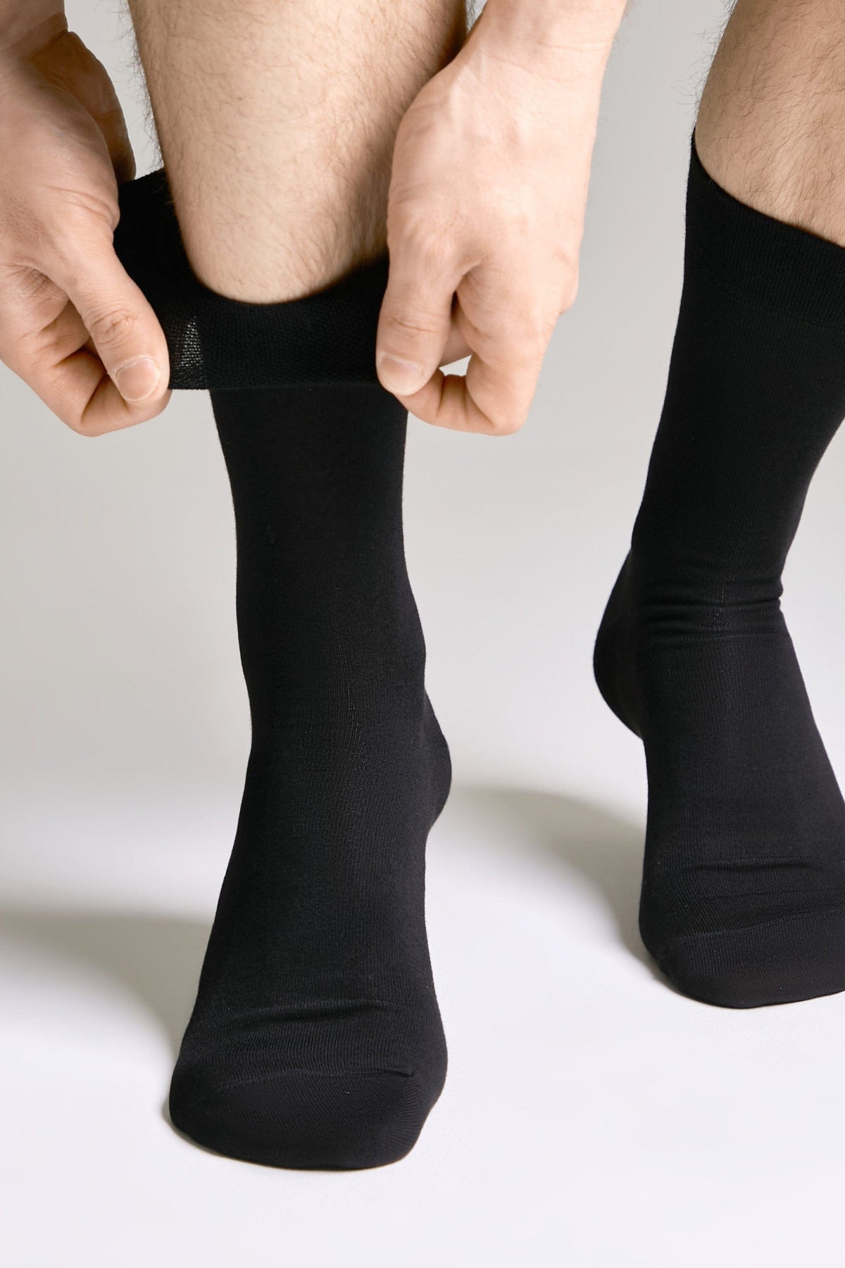 A pair of black crew-length socks being worn by a person, with hands adjusting the cuff of the left sock against a plain light gray background.