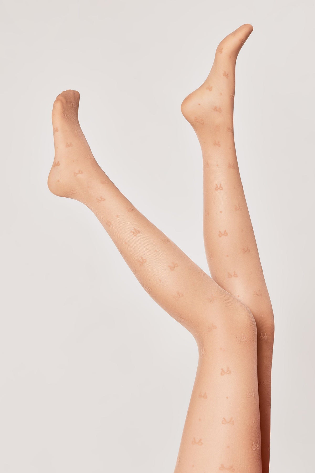 Sheer nude-toned tights with a repeating pattern of small bows are modeled on a pair of raised legs against a plain light gray background.