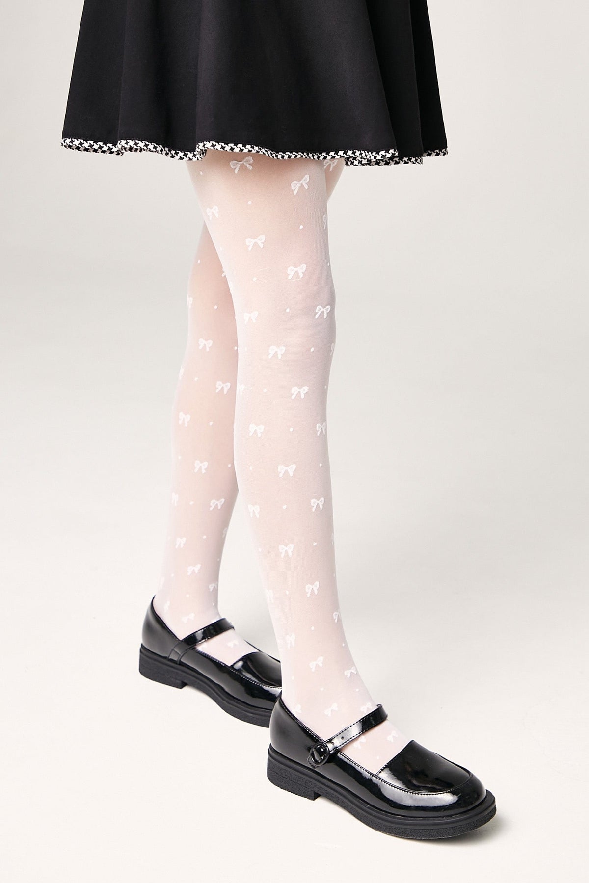 A person wears sheer white tights featuring a repeating pattern of small bows paired with black patent leather Mary Jane shoes and a black skirt.