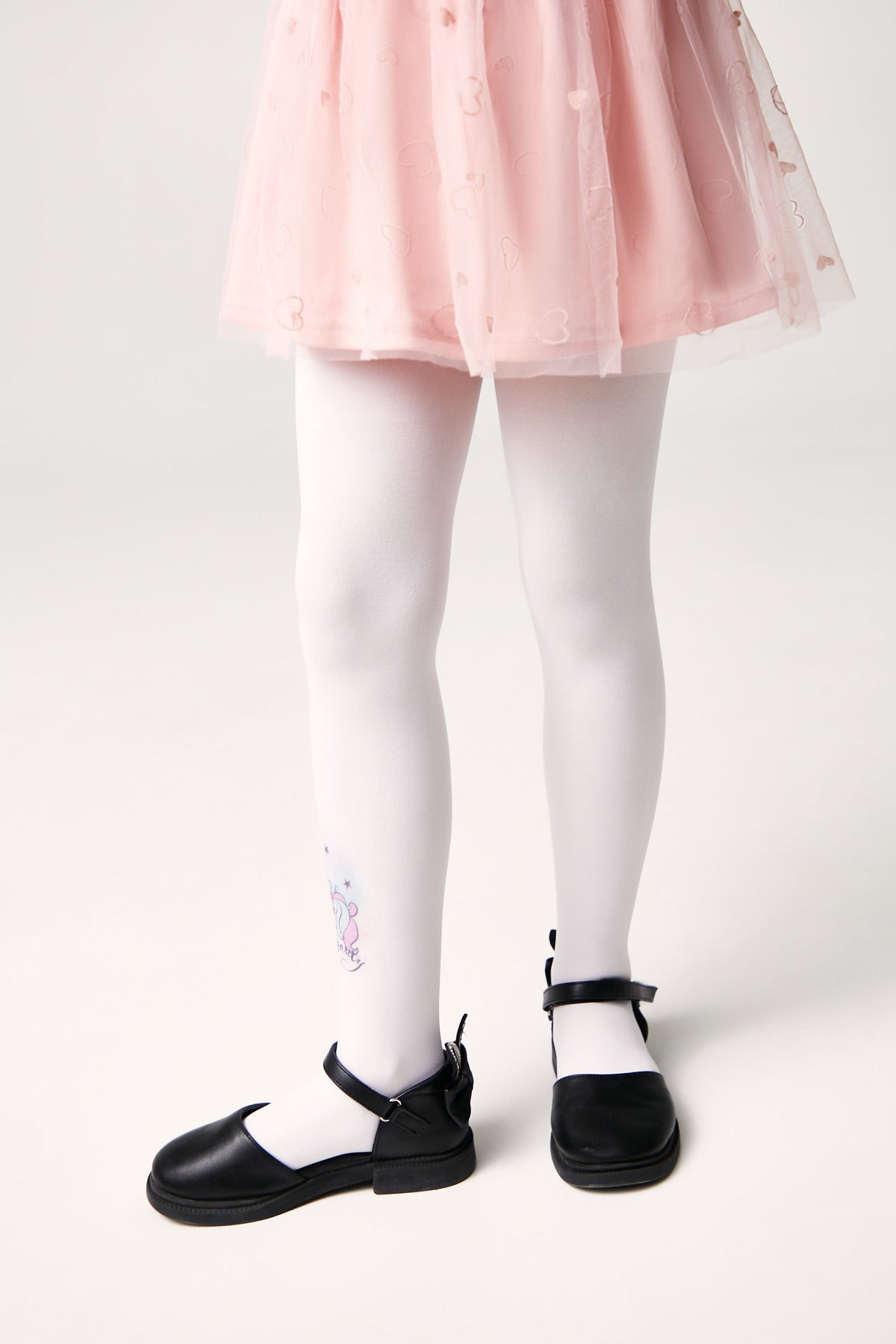 A pair of white tights featuring a small unicorn graphic on the lower leg, worn with a pink skirt and black buckled shoes.