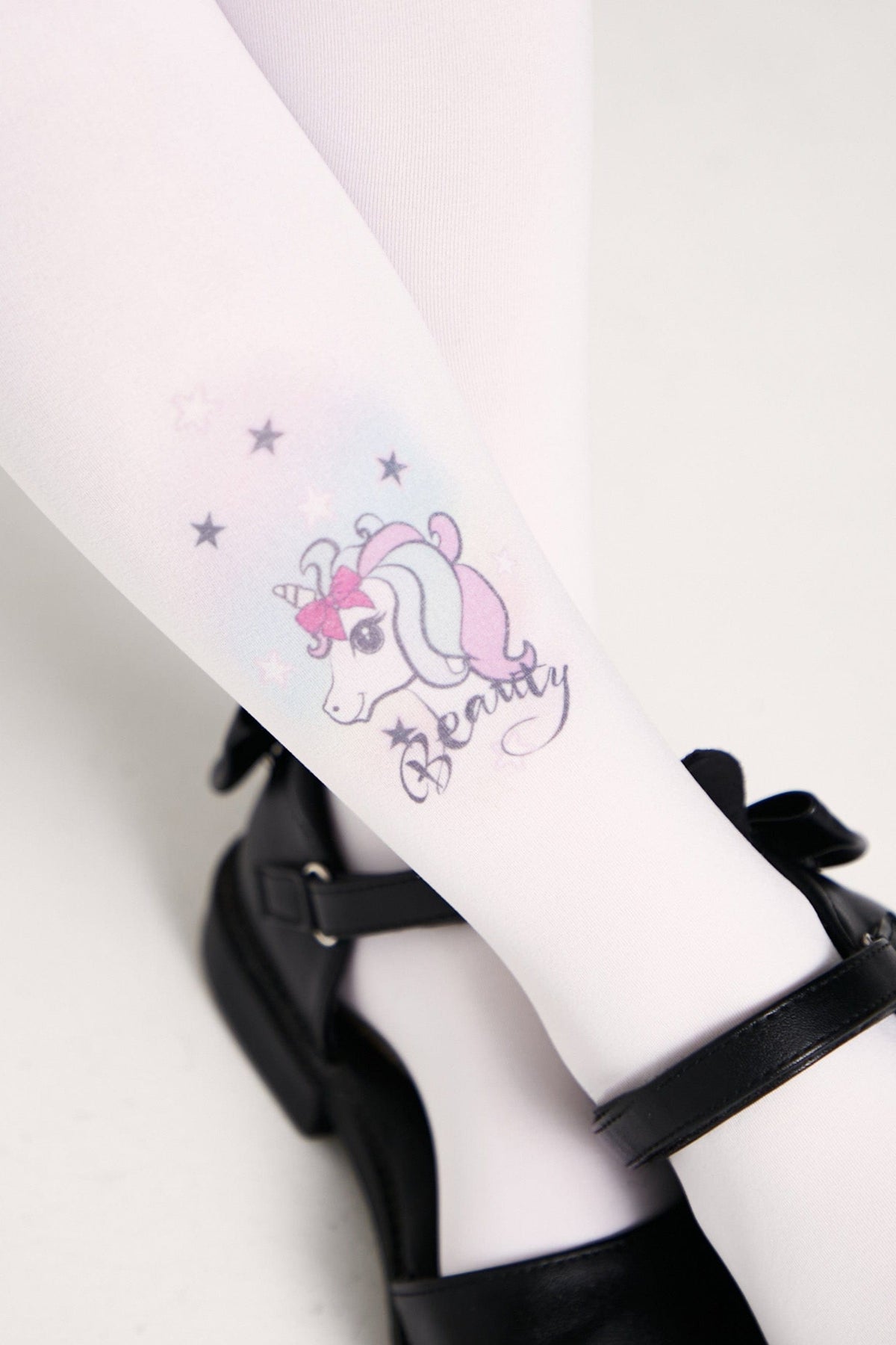 White tights featuring a pastel unicorn graphic with a bow, star motifs, and the word Beauty printed on the calf, paired with black buckled shoes.