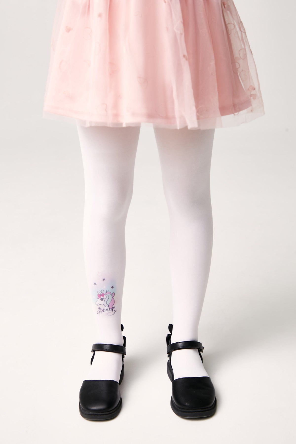A pair of white opaque children's tights with a small unicorn illustration on the right calf, paired with a pale pink tulle skirt and black Mary Jane shoes.