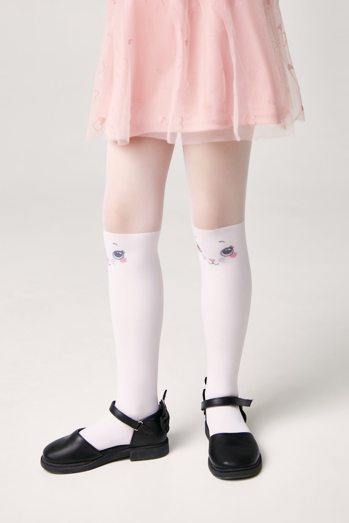 A pair of white tights for a child, featuring a cute cat face design on the knees, worn with a pink skirt and black flat Mary Jane shoes.