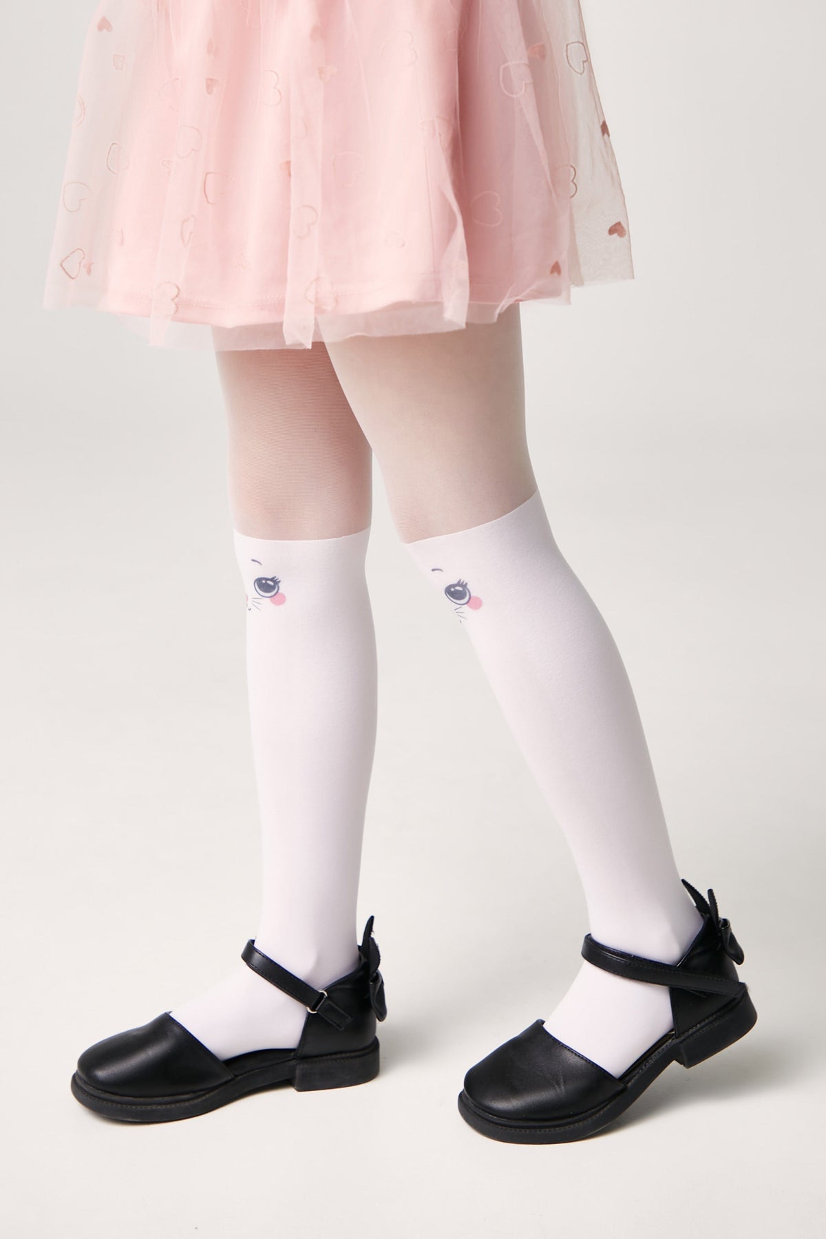 A child wearing white knee-high socks featuring a cute cat face design, paired with a pink tulle skirt and black Mary Jane shoes against a plain light background.