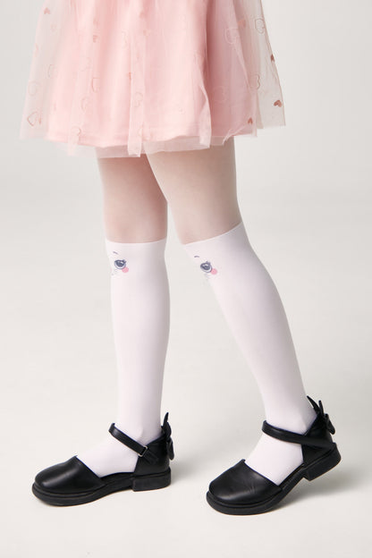 A child wearing white knee-high socks featuring a cute cat face design, paired with a pink tulle skirt and black Mary Jane shoes against a plain light background.