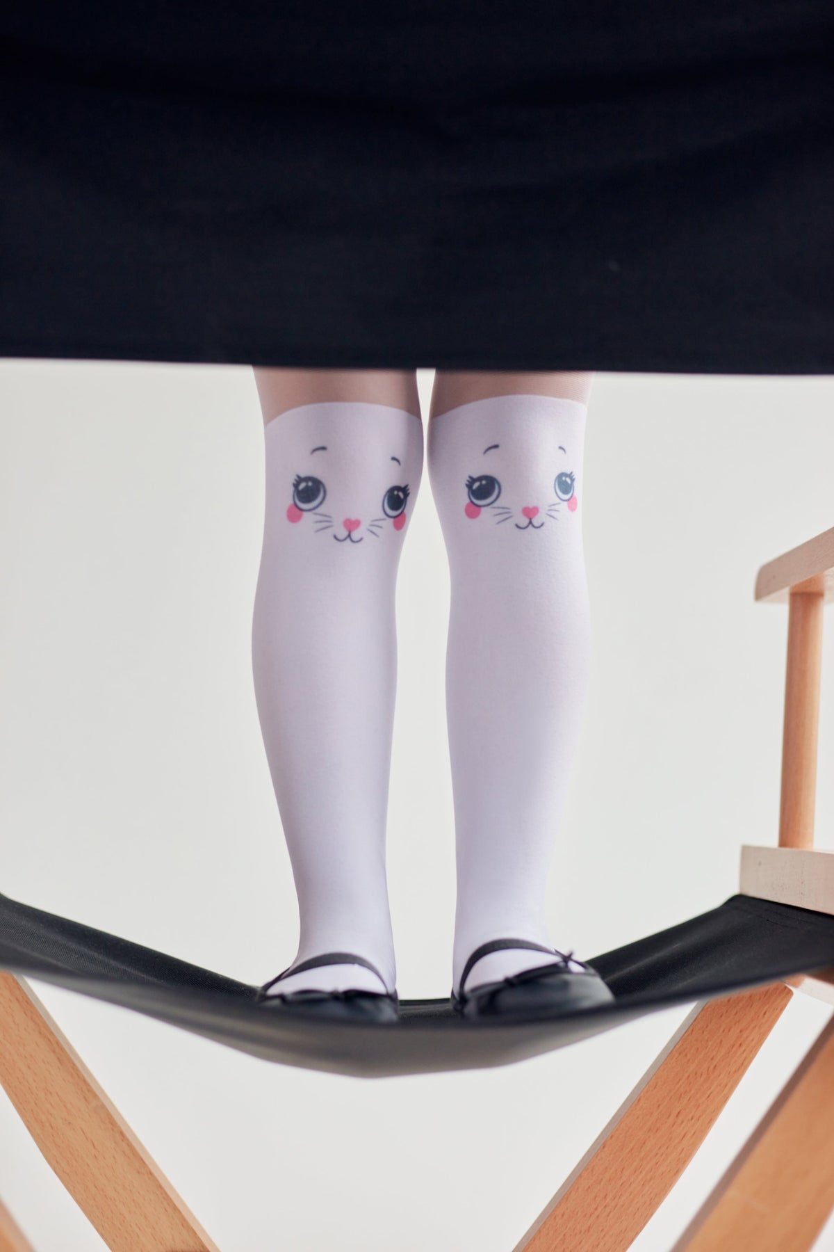 White knee-high socks with a cute cat face print on each knee, worn with black ballet flats while standing on a wooden director chair.