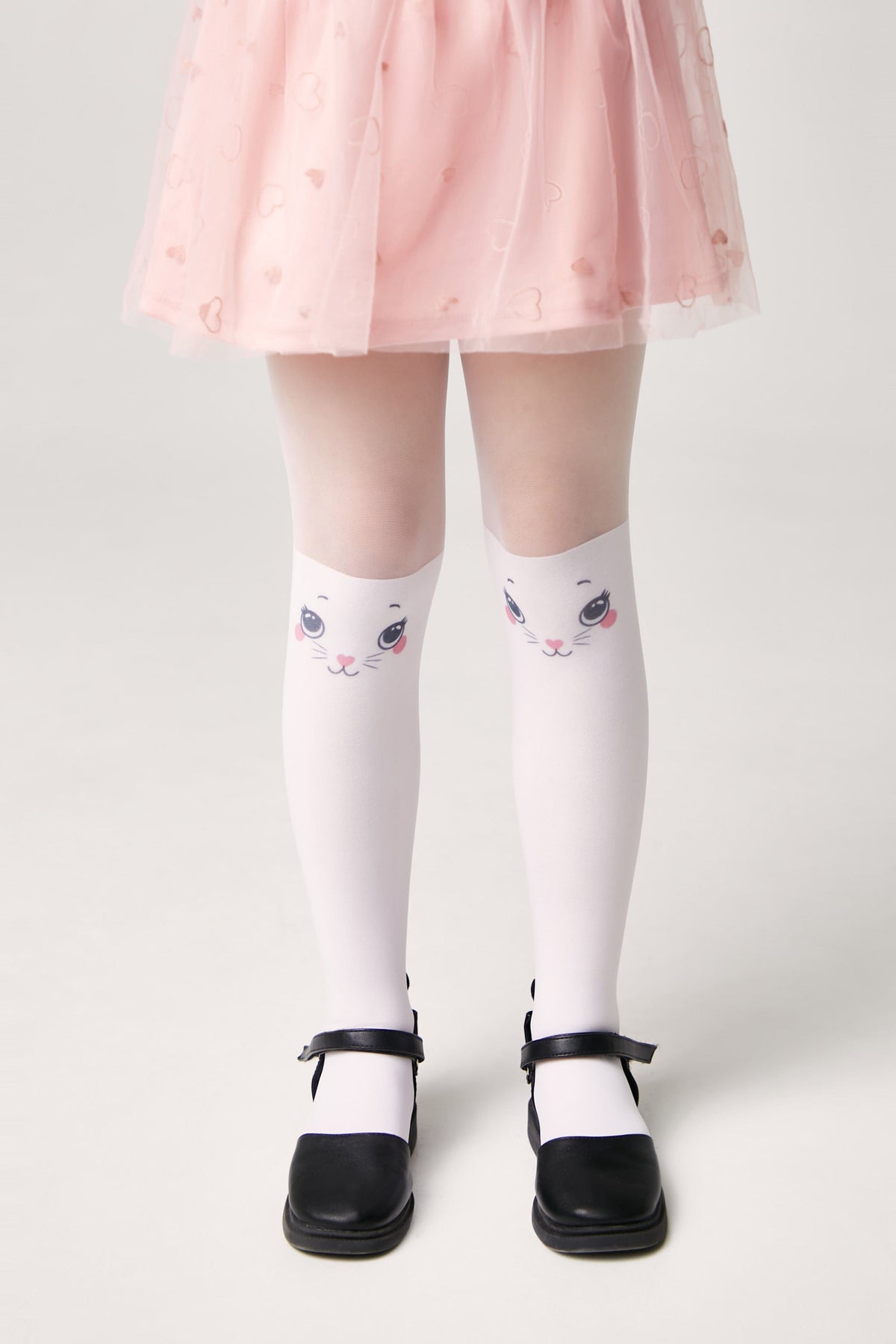 A pair of white tights featuring a cartoon cat face pattern on the knees, worn by a child with a pink skirt and black mary jane shoes.