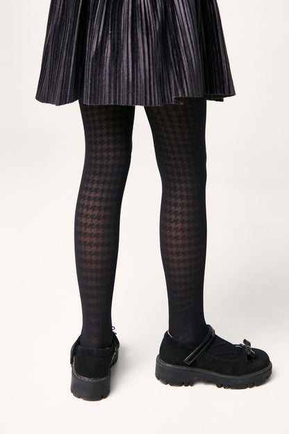 A pair of black opaque tights with a subtle houndstooth pattern worn with a black pleated skirt and black Mary Jane shoes with thick rubber soles.