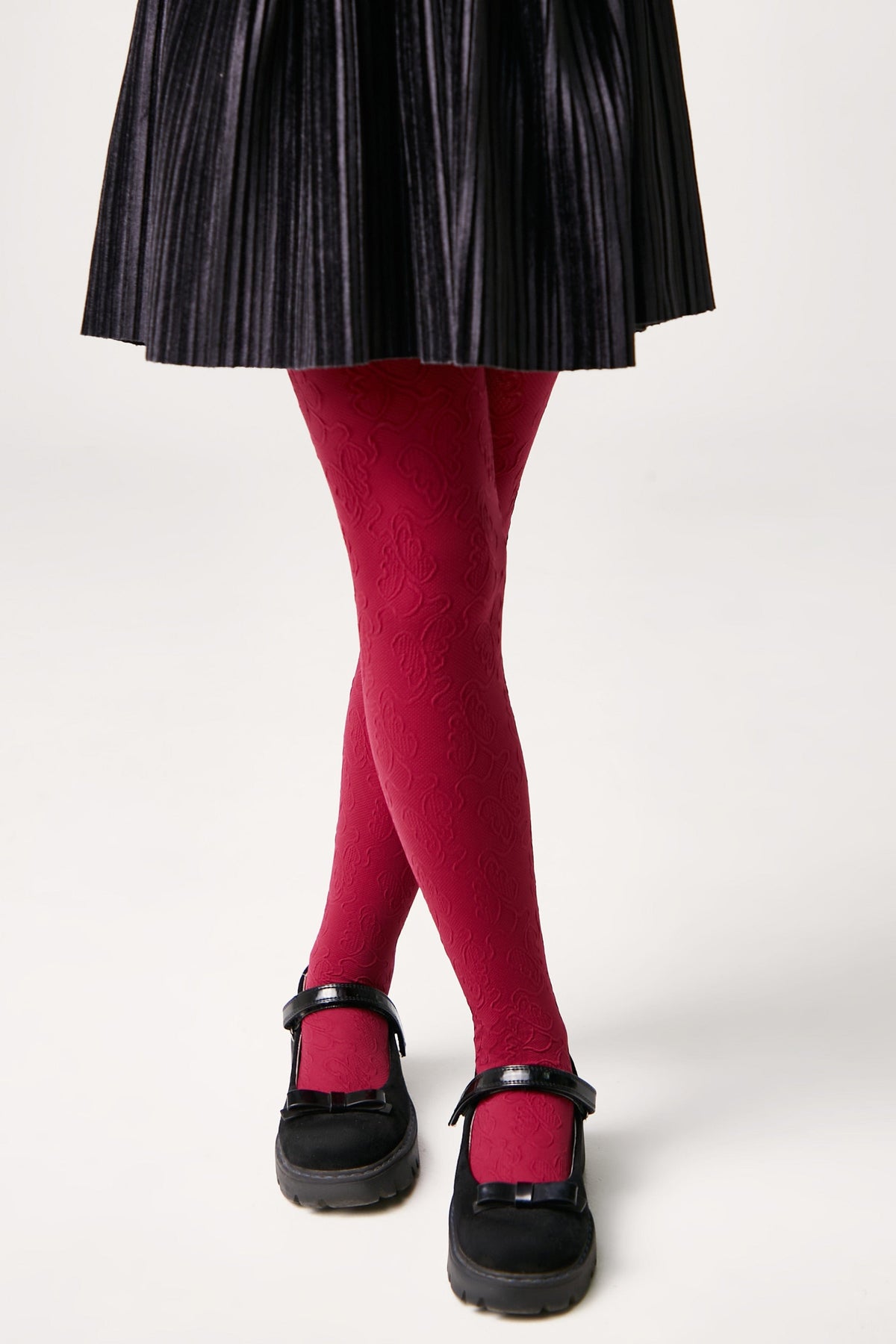 A person wears red patterned tights paired with a black pleated skirt and black Mary Jane shoes against a white background.
