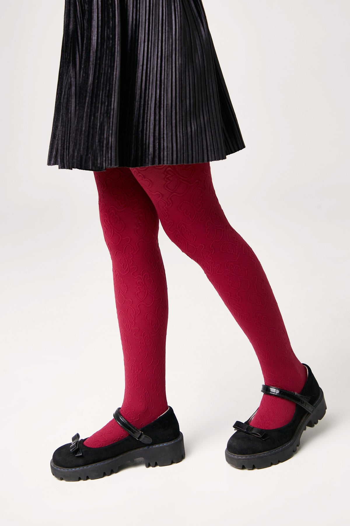 Burgundy tights with a subtle rose-patterned texture worn with a black pleated skirt and black Mary Jane shoes featuring a bow detail on a white background.
