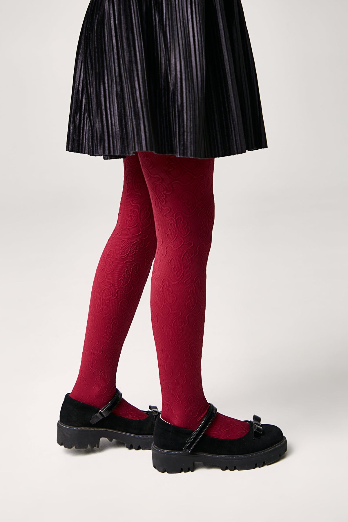 A pair of red patterned tights with a floral-like design, worn with black pleated skirt and black buckled mary jane shoes.