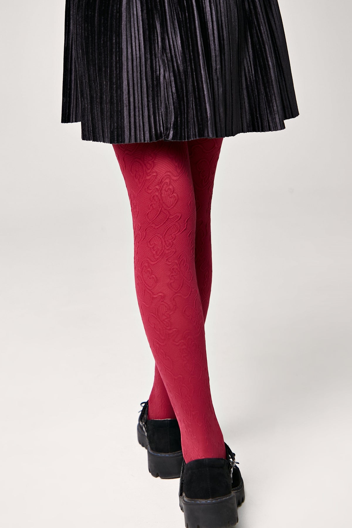 Red patterned tights worn with a black pleated skirt and black platform shoes, viewed from behind against a plain white background.
