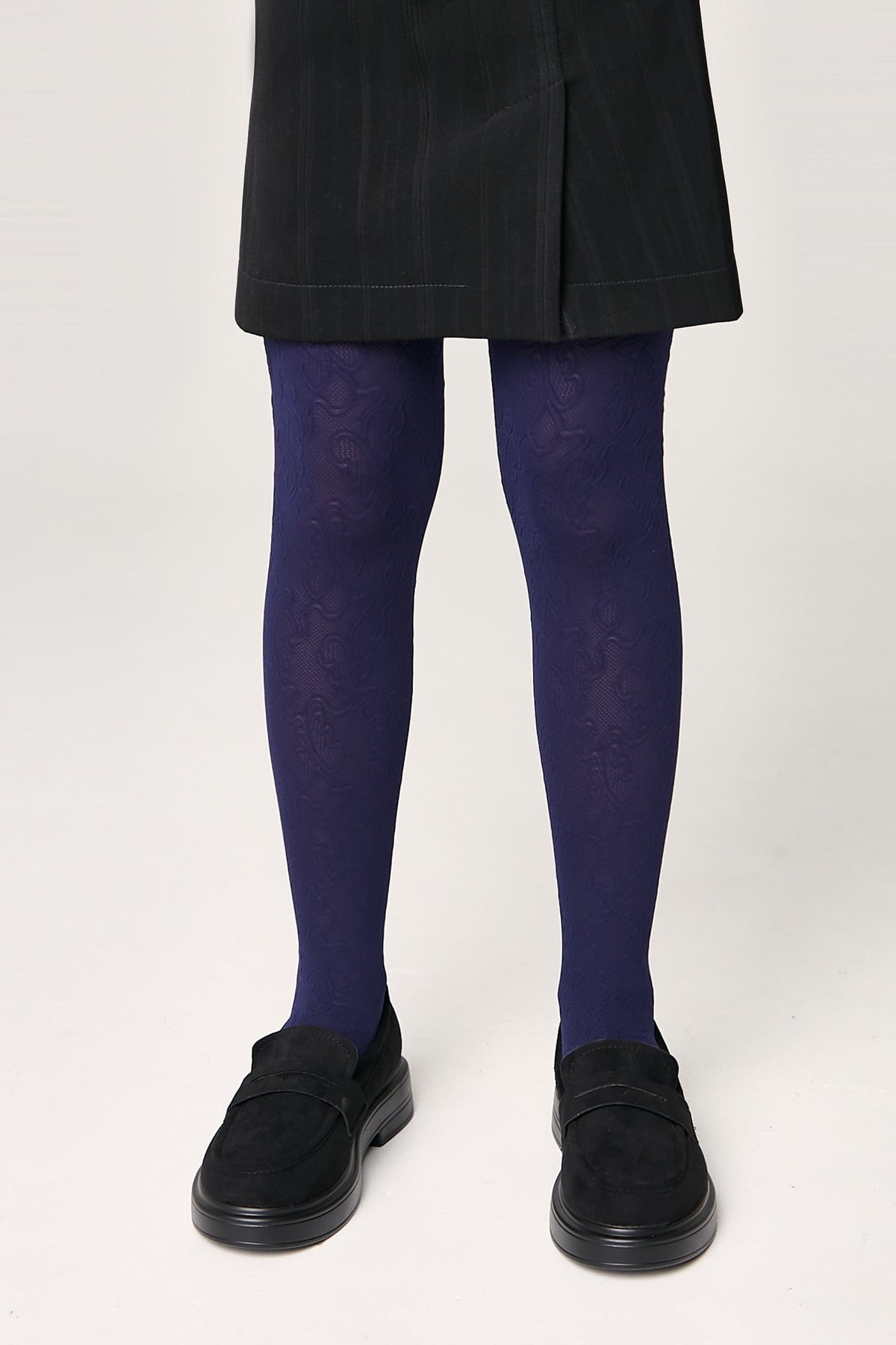 A person wears deep purple tights with an intricate tonal swirl pattern paired with a black skirt and black suede-like loafers.