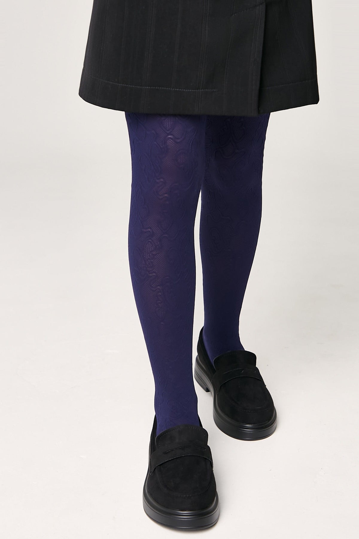 Dark purple patterned tights worn with a black skirt and black suede loafers against a plain white background.