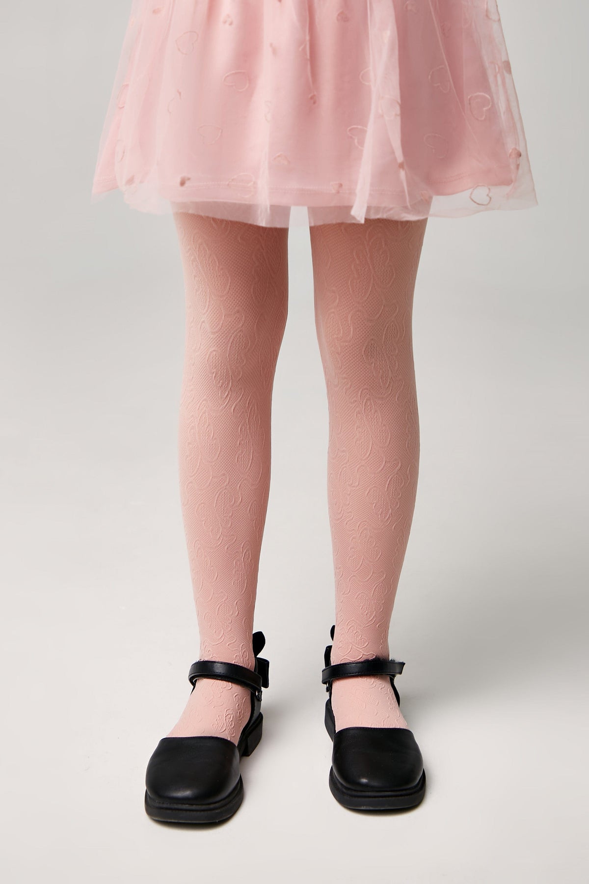 A pair of light pink patterned tights worn with a pink skirt and black flat shoes on a plain grey background.
