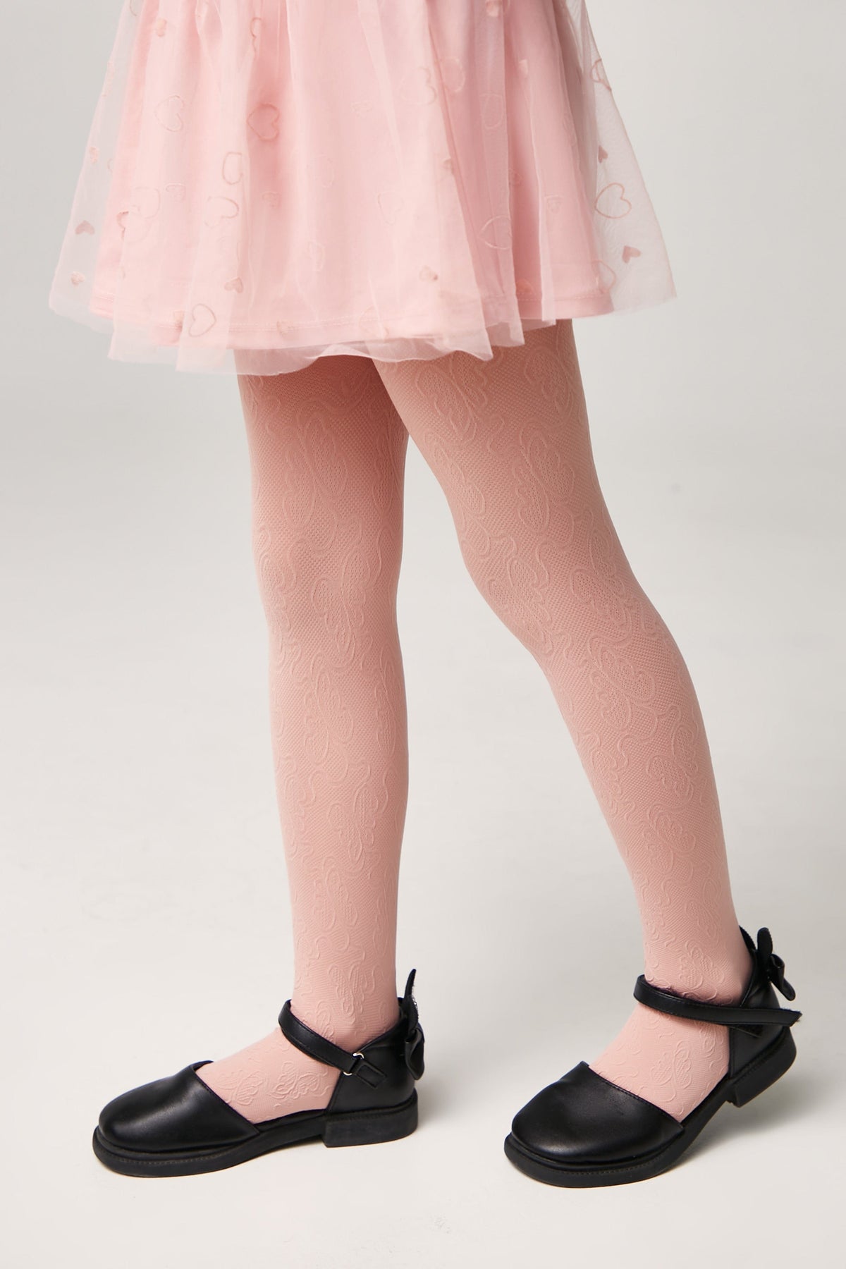 A child wears patterned light pink tights underneath a pink skirt with heart detailing, paired with black buckled flats.