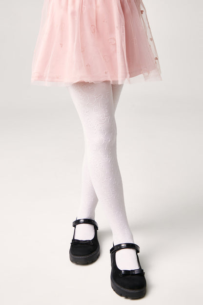 A child wearing white patterned tights and black Mary Jane shoes with small bows, paired with a light pink skirt featuring a subtle heart print.