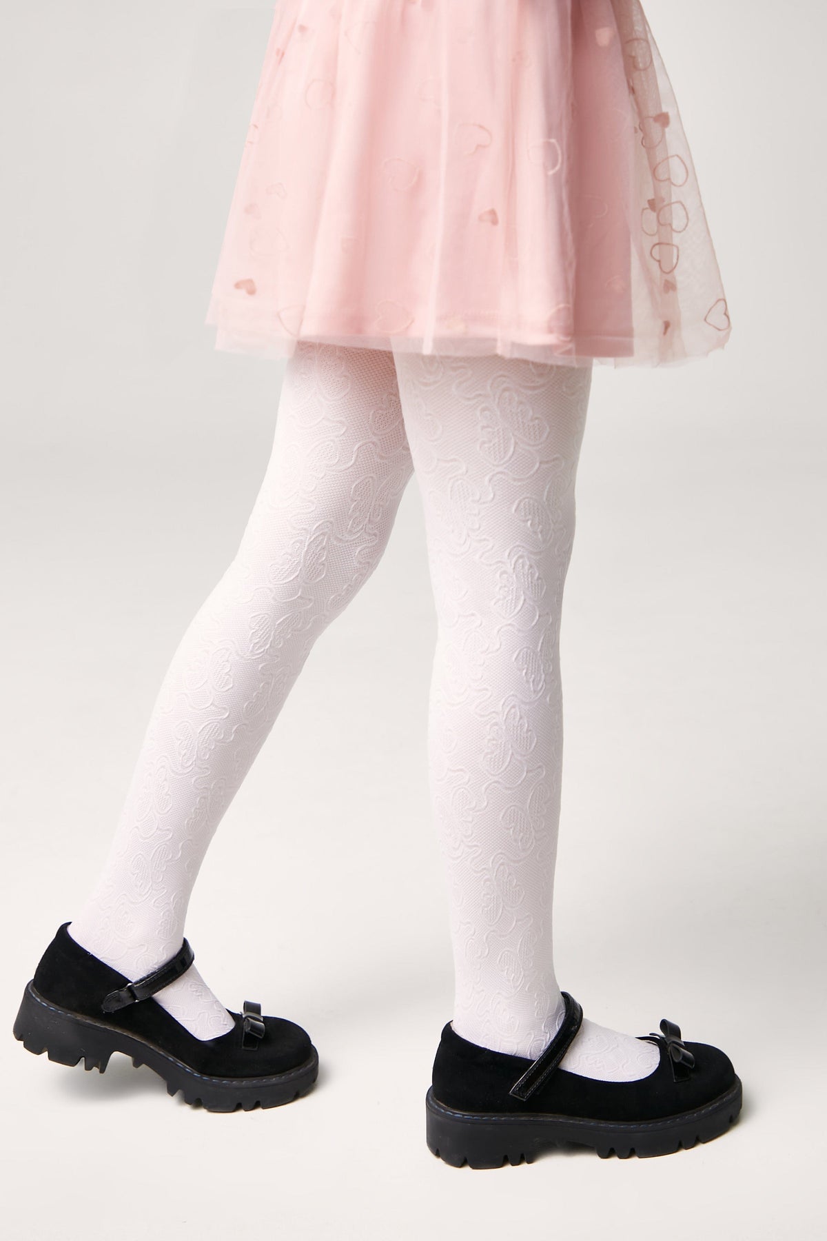 A pair of white patterned tights worn under a light pink skirt with black buckled shoes featuring small bows.